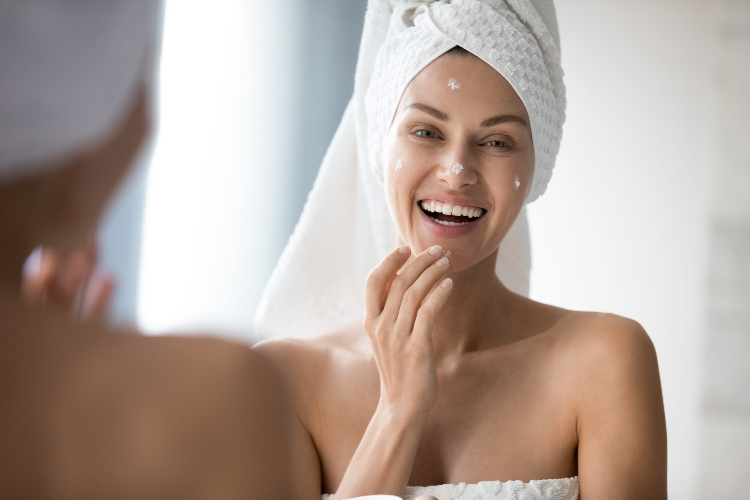 Woman in towel smiling while looking in mirror putting on moisturizer after microneedling in Jacksonville